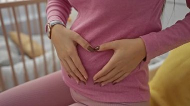 Yatak odasında pembe elbiseli hamile bir kadın. Elleri karnında, tahtadan bir beşiğin yanında. Annelik bağını ve rahat yuvasını aydınlatıyor.