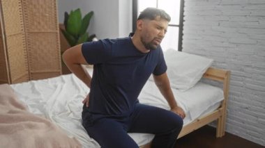 Yatakta sırtı ağrılı genç bir adam, modern bir yatak odasında rahatsızlık gösteriyor doğal ışık ve minimalist dekorla..