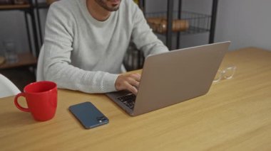 Ofisteki genç adam laptopta işten sonra telefon ve kırmızı bardakla masada dinleniyor. Kapalı alanda tamamlanma ve memnuniyet hissi veriyor.