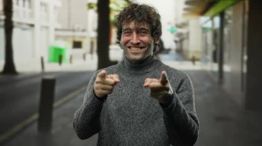 Genç adam gülerek ve parmaklarını şakayla göstererek gündüz vakti bir şehir caddesinde dikilirken neşeli bir şehir sahnesini karizması ve gündelik tarzıyla yakalıyor..