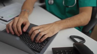 Yeşil önlüklü ve steteskoplu genç erkek doktor modern bir hastane kliniğinde dizüstü bilgisayar üzerinde çalışıyor.