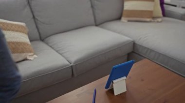 Genç adam rahat oturma odasında tablet kullanıyor, rahat bir kanepe giyiyor, rahat kot pantolon ve beyaz tişört giyiyor, renkli yastıklarla modern iç mekan ve apartman dairesinde ahşap masa.