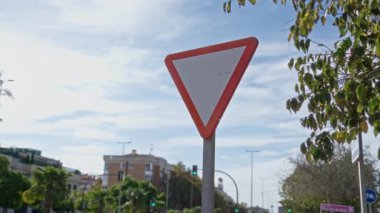 Açık mavi gökyüzü altında yeşil ağaçlar ve binalarla çevrili güneşli bir şehir sokağında trafik üçgeni işareti açık şehir yaşamını aydınlatıyor..