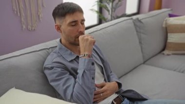 Evdeki kanepede oturan genç adam, modern oturma odasının iç kısımlarında keyifsiz görünüyor. Gün boyunca rahat bir apartman dairesinde tefekkür ederken tarafsız bir ifade takınıyor..