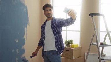 Oturma odasında tekerlekli sandalyeyle duvar boyayan, etrafı kutular ve merdivenlerle çevrili selfie çeken genç bir adam, bir ev yenileme projesi sergiliyor..