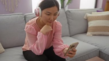 Oturma odasında kulaklık takan akıllı telefondan video sohbeti yapan bir kadın..