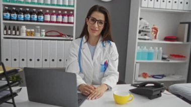 Hastane muayenehanesindeki kadın doktor kollarını kaldırmış, gözlük, beyaz önlük ve steteskop takmış, etrafı tıbbi malzemelerle çevrili ve masanın üstünde dizüstü bilgisayarla kutlama yapıyor..