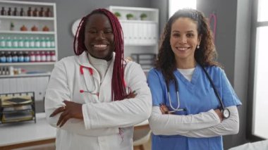 İki gülümseyen kadın doktor, hastane kliniğinde kollarını kavuşturup, sağlık hizmetlerinde profesyonellik gösteriyorlar..