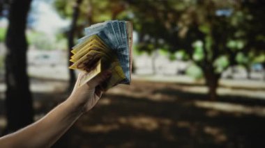 Macar banknotlarını bir park ortamında elinde tutan adam, arka planda bulanık ağaçlarla doğal bir ortamda para ve finans sergiliyor..