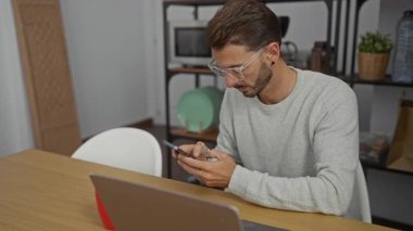 Modern ofisteki genç adam, bilgisayarını ahşap bir masanın üzerine açıp, yoğun bir çalışma ortamını gösteren akıllı telefonlara odaklanıyor..