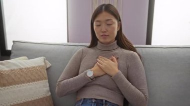 Oturma odasında, elleri göğsünde, rahat bir evde, kanepede oturan, süveter ve saat giyen, konforu kucaklayan, barışçıl Asyalı güzelliği yansıtan bir kadın..