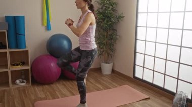 Spor kıyafetleriyle spor yapan bir kadın spor yapıyor spor minderinde spor yapıyor spor topları ve kapalı alanda parlak pencere ışığı eşliğinde yoga malzemeleri sergiliyor.