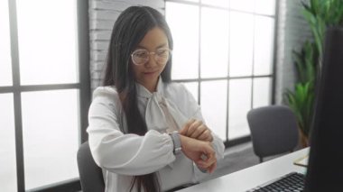 Ofisin içindeki masaya gülümseyen gözlüklü bir kadın modern işyerinde saati kontrol ediyor doğal ışık ve yeşillik etrafında sakin bir ortam yaratıyor.