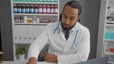 Sakallı, gözlüklü ve beyaz önlüklü bir adam klinikte çalışıyor. Tıbbi malzemeler ve ofis dosyalarıyla çevrili ekipmanları inceliyor..