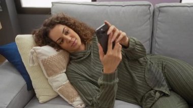 Şık yastıkları ve rahat kıyafetleri olan, rahat bir oturma odasında akıllı telefon kullanan bir kadın, boş bir anı içeride yakalıyor..