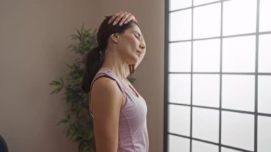 Spor salonunda boynunu uzatan, camdan içeri güneş ışığı süzülen, atletizm tankı takan ve spor topunun yanında gevşemiş olarak görünen bir kadın..