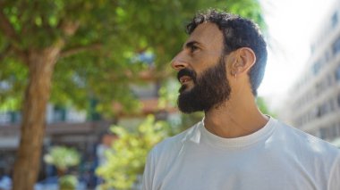 Sakallı genç bir adam şehir parkında duruyor.