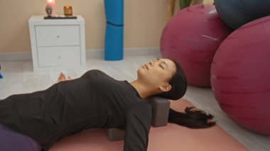 Çinli bir kadın içeride rahatlama egzersizi yapıyor spor salonundaki yoga minderinde egzersiz topları ve yakınlarda bir meditasyon seti ile yatıyor..