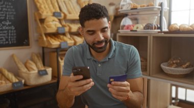 Etrafı ekmek ve hamur işleriyle çevrili bir pastanede telefon ve kredi kartı kullanan İspanyol genç adam.
