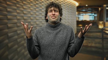 Bir otel resepsiyonunda kafası karışmış görünen, süveter giyen, yakışıklı görünümlü genç bir adam modern bir iç mekan izlenimi uyandırıyor, düşüncelere dalmış çekici bir İspanyol yetişkini yakalıyor..