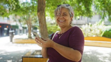 Şehir parkında elinde telefon tutarken gülümseyen bir kadın, yemyeşil ve canlı bir arka planı olan bir şehir ortamında yakalandı..