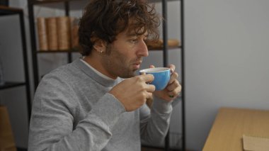 Kitap rafları ve arka planda asgari dekor olan bir ofis ortamında kahveden hoşlanan genç bir adam, sıradan ve odaklanmış bir atmosferi canlandırıyor..