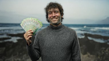 Bir adam, elinde Rus rublesi tutarken gülümsüyor. Sahilde duruyor, açık hava finansal başarısının ve eğlencenin neşeli ve kaygısız özünü yakalıyor..