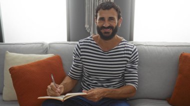 Oturma odasındaki koltukta oturan sakallı gülümseyen genç adam bir deftere yazı yazarak evde rahat bir anı yakalıyor..