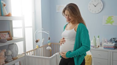 Bebek odasında gülümseyen hamile bir kadın kucağında bebek oyuncakları ve bebek malzemeleriyle dolu sıcak ve neşe yansıtan yumuşak gün ışığı altında.
