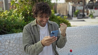 Güneşli bir günde şehir parkında başarıyı akıllı telefonla kutlayan genç adam, yeşillik ve şehir çevresi arasında heyecanını dile getiriyor..