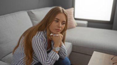 Genç bir kadın, düşünceli bir ifadeyle oturma odasında oturuyor, uzun sarı saçlar sergiliyor, güzel bir yüz, çizgili bir gömlek ve saat giyiyor, rahat bir evin içini yansıtıyor.