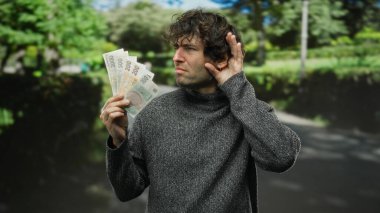 İspanyol asıllı bir adam elinde bir park ortamında dinleme jesti ile Czech Koruna banknotları tutuyor. Merakını doğal çevreyle harmanlıyor..