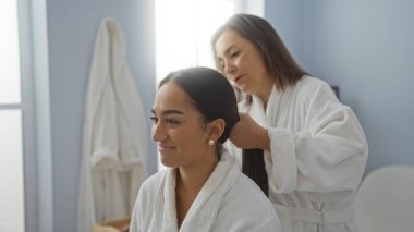 Anne ve kızı bir kaplıca merkezinde bornoz giyip birbirlerine bağlanırken anne, kızının saçını kapalı bir güzellik salonunda sevgiyle bağlıyor.