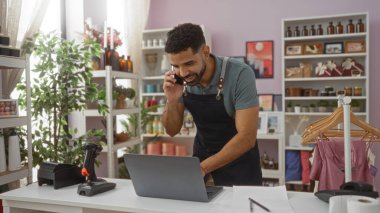 Ev dekorasyonu mağazasında çalışan genç bir adam dizüstü bilgisayar kullanırken telefonla konuşuyor. Etrafı dekoratif eşyalar ve bitkilerle çevrili.