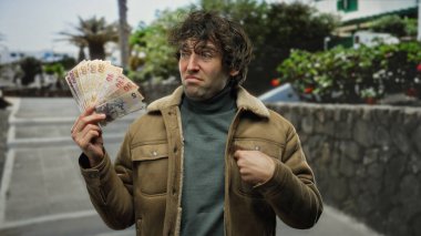 Elinde Türk lirası tutan İspanyol bir adam şüpheli bir ifadeyle şehir sokaklarında finansman ve kentsel çevre temalarını öne sürüyor..