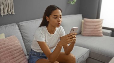 Modern bir oturma odasındaki kanepede oturan genç bir kadın akıllı telefonunu kullanıyor.