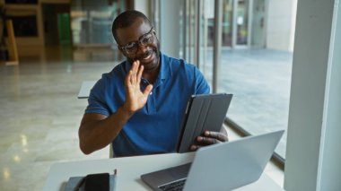 Ofisteki yakışıklı siyah adam bir video görüşmesi sırasında gülümsüyor ve tablete el sallıyor.