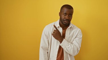 Sarı duvar arkaplanının üzerinde duran yakışıklı Afrikalı genç adam, boşluğu kopyalamak için parmağıyla işaret ediyor.