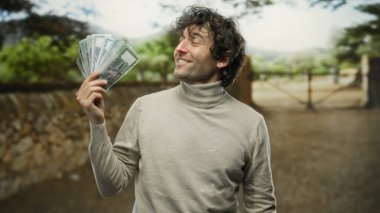 Parkta elinde bangladeş banknotları tutan İspanyol bir adam arka planda ağaçlarla gülümseyen, finansal başarı ve mutluluğu yakalayan bir adam..