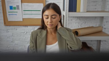 Ofisteki genç bir kadın çalışırken boyun ağrısı çekiyor. Etrafı çizelge ve dosyalarla çevrili. İş stresi senaryosunu yansıtıyor..