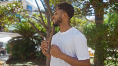 Beyaz gömlekli genç adam telefonunu tutuyor ve arka planda yeşil ağaçlar ve binalarla dolu bir parkta gülümsüyor.