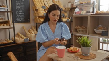 Ekmek ve hamur işleriyle çevrili kahve ve kruvasanın tadını çıkarırken fırın kafede oturan genç bir kadın.