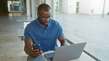 Bir ofiste çalışan genç bir adam dizüstü bilgisayar ve akıllı telefon kullanarak, kapalı bir çalışma alanındaki görevlere odaklanıyor.