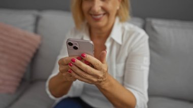 Orta yaşlı, kısa sarı saçlı bir kadın. Evinin oturma odasında akıllı telefon kullanıyor.