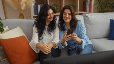 Kadınlar gülümsüyor ve rahat bir oturma odasında akıllı telefon kullanıyor, sıcak bir aile ve kapalı alanda dostluk ortamı sergiliyor..