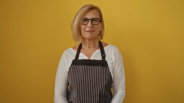 Sarışın, kısa saçlı, gözlüklü, çizgili önlüklü, sarı arka planda izole edilmiş güzel olgun bir kadın.