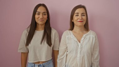 Anne ve kızı pembe bir arka plana karşı birlikte duruyorlar. Sevgi dolu ilişkilerini ve mutlu ifadelerini sergiliyorlar..