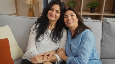 Kız kardeşler rahat bir oturma odasındaki kanepede birlikte oturur, sıcak bir şekilde gülümserler, rahat ve davetkar bir ortamda sevgi ve aile bağları sergilerler..