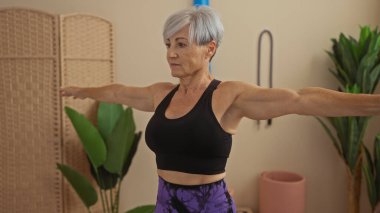 Kısa gri saçlı, spor salonunda egzersiz yapan, siyah bluz ve mor tayt giyen, kollarını uzatan güzel yaşlı bir kadın..