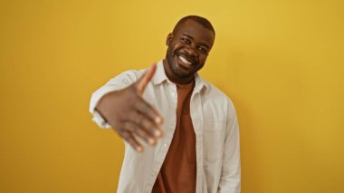 Yakışıklı, Afro-Amerikalı bir adam, izole edilmiş sarı bir arka plana gülümsüyor ve elini uzatıp hoş geldin diyor.